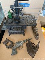 Overview of cast iron toy stove, green glass bottle, fish-shaped piece, revolver toy, bird figure, and toy train car on table.