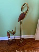 Photo showing the set of three brass and wood stork birds of varying sizes against a green wall and wooden floor.