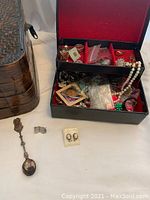 Overview of two-tiered black jewelry box containing assorted jewelry and pearl-like beads, with collectible spoon and APA earrings displayed outside.