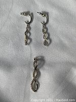 Photo shows pair of white gold twisted leaf design dangle earrings with stud backings and a matching pendant, all laid on a white fabric background.