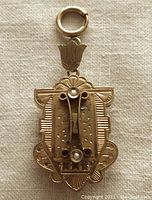 Front view of antique Victorian gold-filled fob locket pendant showing detailed engraved design, fancy floral bail, and two seed pearls.