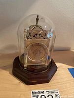 Antique brass table clock in glass dome on wooden base, front view showing clock face with Roman numerals.