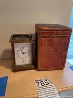 Full view of carriage clock next to its leather case on a wooden surface.