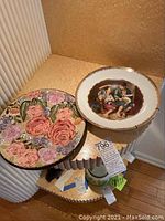 Two collector plates and one ceramic plate shown on wooden table, including floral and boys playing lute images.
