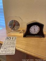Two small table clocks side by side on a wooden surface, one with sterling silver overlay and the other clear octagonal plexiglass.
