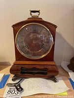 Full view of Warmink carriage clock with wooden case, brass handle, and decorative metal feet, standing on a surface with appraisal document and winding key.