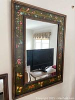 Front view of large rectangular wall mirror with hand-painted floral and bird motif on the wooden frame. Mirror reflects a room interior including a TV screen and window with curtains.