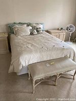 Bedroom bench upholstered in light striped fabric with carved wooden legs and frame, placed at foot of bed with pillows and bedspread visible.