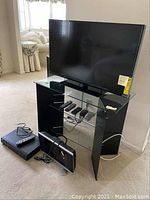 Front view of BellO metal and glass TV stand holding Vizio 38-inch TV, RCA DVD player and Sharp RP 13 TV with remotes on glass shelf.