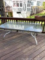 Rectangular patio table with textured glass top and white metal frame on wooden deck, taken from side angle showing four curved legs and overall structure.