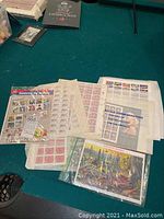 Photo showing multiple sheets and blocks of assorted U.S. postage stamps spread on a green surface, some loose and some in protective sleeves.