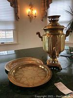 Photo showing the Russian style brass samovar with its handles and spout, along with the two brass trays in the background on a table.