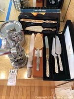 Photo showing three cake server sets including boxed Neiman Marcus set, Silea set, and third set along with metal ice bucket, shaker, and strainer.