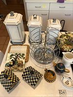 Photo of three white hexagonal kitchen canisters with silver trim and lids, assorted kitchen decor, teapots, pitcher, decorative tile, and metal rack on kitchen counter.