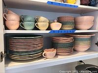 Wide shot of cabinet shelves filled with dinnerware plates, cups, serving bowls and covered sugar bowl in pastel colors with ridged designs.
