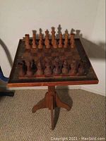 Full view of wooden pedestal chess board with set chess pieces arranged on top, showing front and one side.