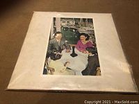 LP cover with four people seated outside at a white table near water and boats