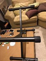 Black Yamaha T-shaped keyboard stand with padded supports shown indoors on a carpeted floor beside a couch and coffee table.