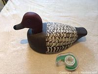 Photo of the wooden duck decoy showing the painted detailed feathers, reddish-brown head, and dark brown tail, placed on a light fabric background with a tape dispenser nearby for scale.