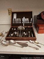 Open wooden presentation box showing top lid with butter knives and bottom tray with spoons, forks and ladles