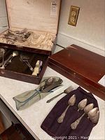 Photo showing multiple pieces of silverware including spoons and forks in vintage wooden boxes and cloth wraps on a counter.