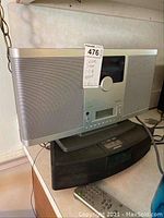 Front and left side view of silver Brookstone radio showing speaker mesh, control panel with large knob, screen, and buttons. Black Bose radio is below it with cords visible.