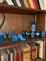 Shelf view of blue vintage glass items including decanter, boot, inkwell, vase, and glasses displayed alongside books.
