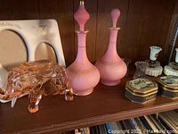 Pink glass elephant ashtray with two pink glass decorative vases with stoppers, decorative tins and ceramic anniversary frame on wood shelf