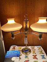 Full view of the vintage converted oil lamp with two white milk glass shades, set against a wood paneling background on a butterfly-patterned surface.