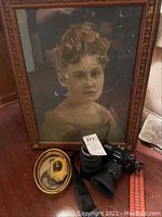 Large framed vintage photographic portrait of a woman with curly hair in a decorative wood frame, positioned behind a Canon camera and two smaller framed portraits on a wooden surface.