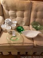 Overall lot on couch showing all glass items including green bowls, white glass bowl shaped like a shell, white cups, clear pedestal bowl, and white pitcher.