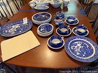 Blue and white china pieces laid out on a wooden table, including plates, platters, multiple cups with saucers, a creamer and sugar bowl, and tower dishes with the Willow pattern decoration.