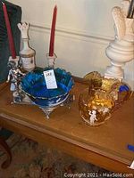 View of the full lot showing the blue glass bowl on silver plated base with cherub, gold glass pitcher, and candle holder with candle on wooden table.