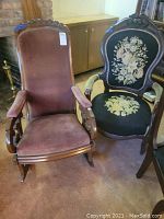 Front view of two vintage chairs side by side, a brown velvet rocking chair with carved wooden top and armrests, and a black floral needlepoint armchair.