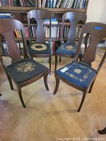 Four vintage wooden chairs with carved backs and floral needlepoint seat covers placed on carpeted floor in front of bookshelf.