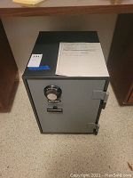 Full front view of a gray FireFyter safe sitting on a floor with a combination dial lock and a handle on the door, paperwork on top.