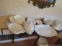 Wide view of the entire white stoneware dinnerware set arranged on a wooden table including plates, bowls, cups, and covered serving dish.