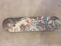 Top view of skateboard deck showing detailed graphic artwork with skulls and abstract figures on worn wooden surface.