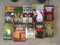 Image showing 10 Stephen King novels arranged on floor, including 'Cujo', 'The Green Mile', 'It', 'Pet Sematary', 'The Dark Half', 'The Tommyknockers', 'The Gunslinger', worn book, 'Misery', and 'The Long Walk'.