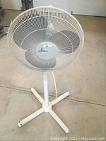 Front angle of the white Ocean Breeze stand up rotating fan showing protective metal grill, three blades, and three-legged base stand on a concrete floor.
