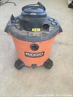 Front angle view of Ridgid orange/black shop vac with power cord and caster wheels, showing RIDGID branding and connection openings, no hose.