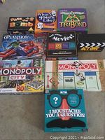 Photo showing all 9 board games arranged on floor, titles clearly visible including Monopoly City, Monopoly, Operation, Pass the Bomb, Tribond, Mad About Movies, What's Your Like?, Act One, and I Moustache You a Question.