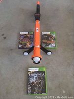 Top down image showing orange and black Xbox 360 gun controller with three Xbox 360 Cabela's hunting game cases around it on floor.