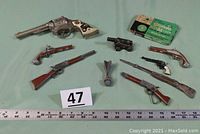 Full spread of vintage cap guns arranged on a cloth with ruler for size reference including the Mattel Greenie Stik-M-Caps box