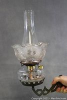 Side view of the assembled kerosene oil lamp held in hand, showing clear glass chimney, decorative glass shade with ruffled edge, glass font with brass components, and cast iron bracket with swivel mechanism.