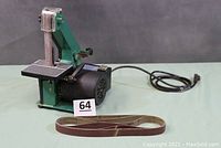 Front view showing the belt sander mounted with belt and connected power cord, with one sanding belt laid in front