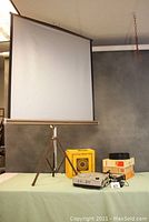 View showing the assembled projection screen on tripod stand and the projector, boxes and trays on the table.