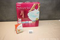 Photo of the boxed Homedics Pedicure Spa and a small box of Baby Foot exfoliate peel on table with number card
