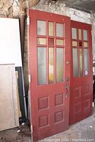 Two red painted church doors leaning against wall with visible hinges and door hardware.