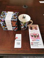 Porcelain mug with colorful Indigenous bear-inspired artwork sitting on wooden table next to matching box and auction brochures.